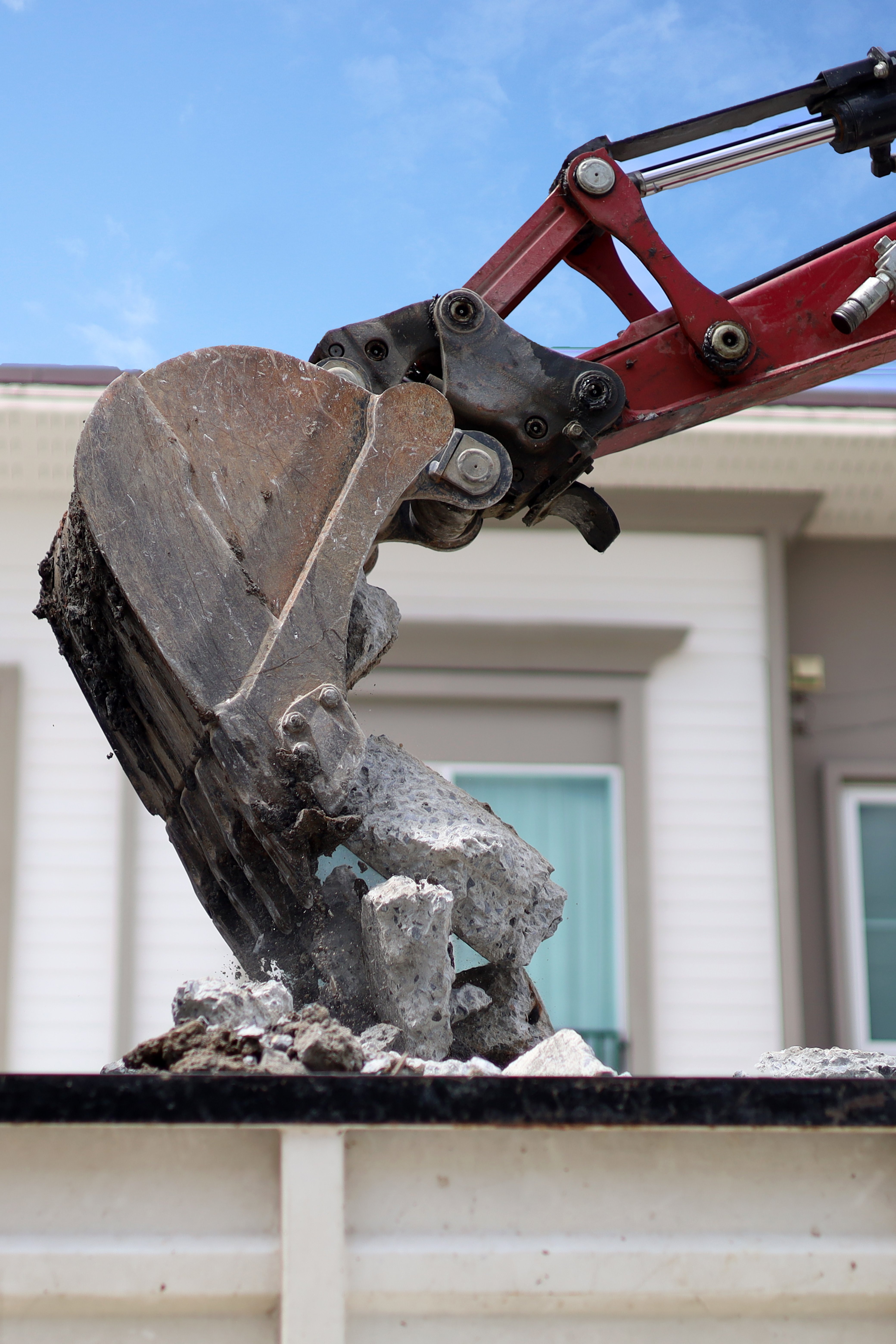 A close-up shot of a red excavator bucket lifting a large chunk of concrete debris into the air.